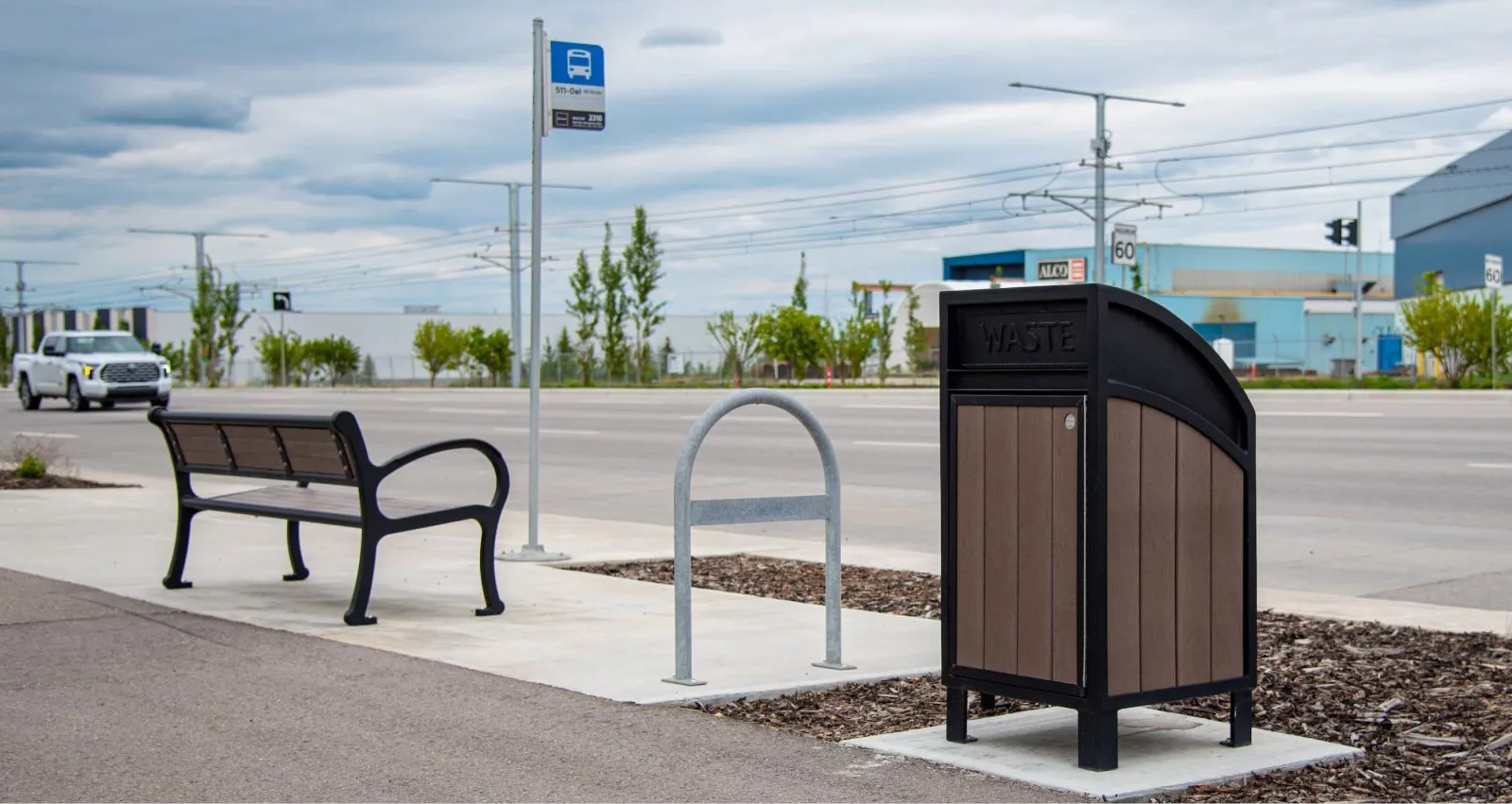 The Mountain Classic bench and Modena Curved Top waste receptacle provide functional seating and waste disposal at a bus stop. The bench features a black frame and recycled plastic lumber, while the waste receptacle has a brown lumber and black curved top.