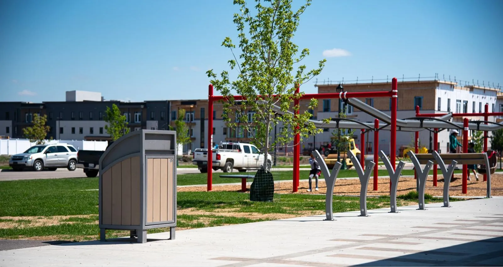The Modena Curved Top Waste Receptacle is shown in a park setting, providing a convenient and stylish waste disposal solution. The bike rack is visible in the background.