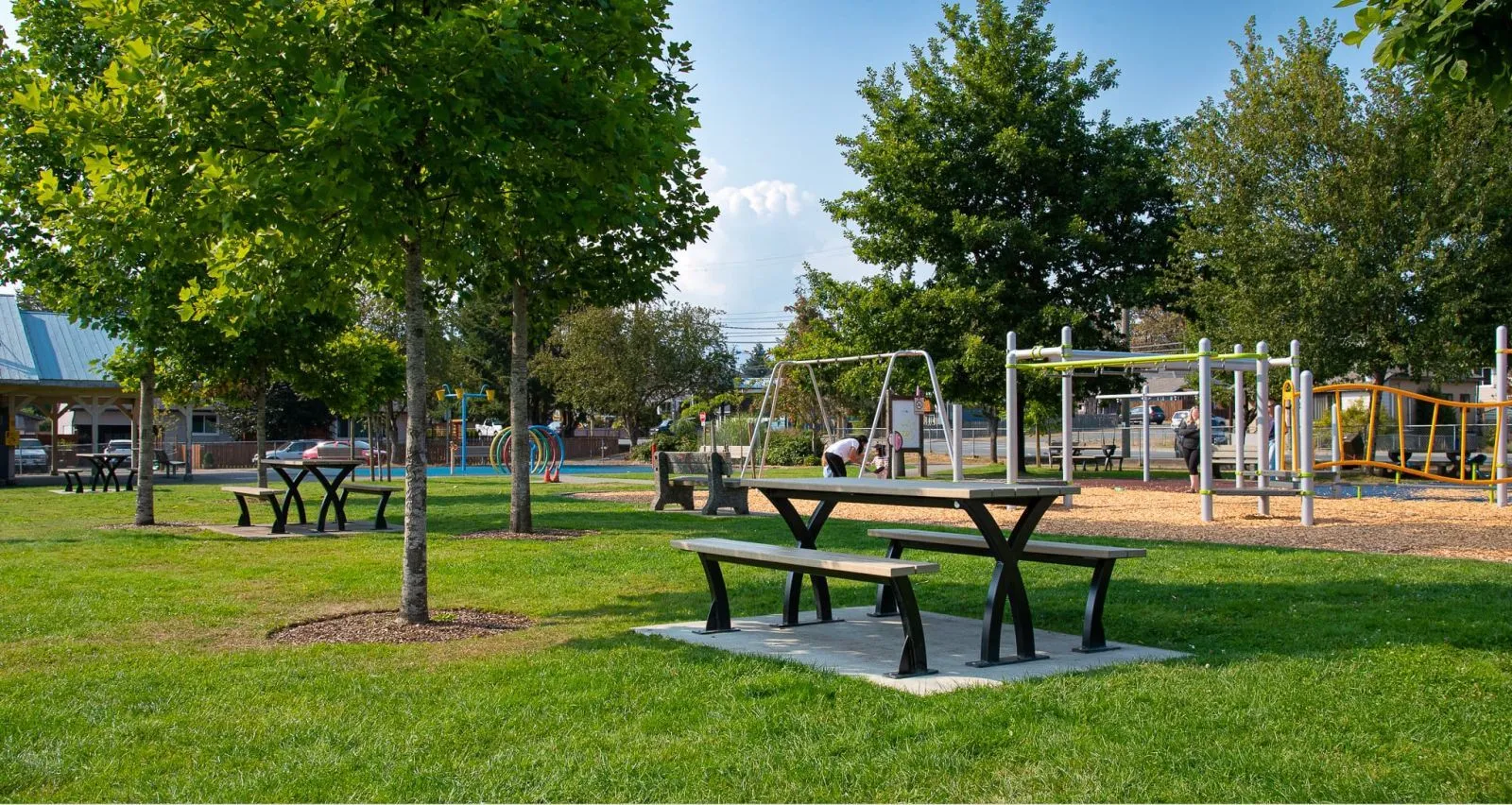 Several picnic tables and benches are arranged in a grassy park area with playground equipment. The tables feature a unique X-shaped leg design and light-colored tops.