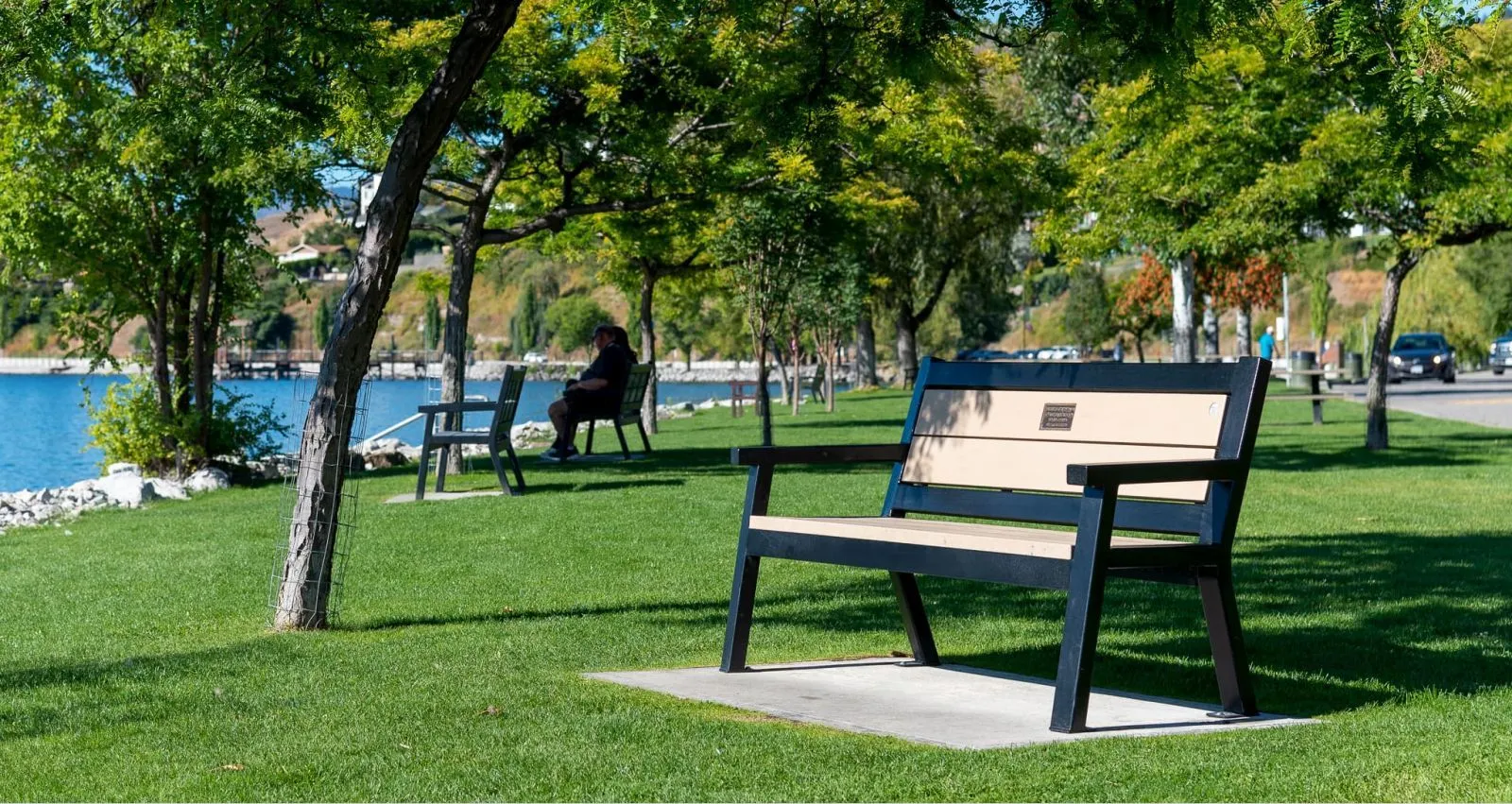 Angled leg memorial bench installed in a lakeside park. The bench features recycled plastic lumber and a black powder-coated frame, providing a durable and comfortable seating option.