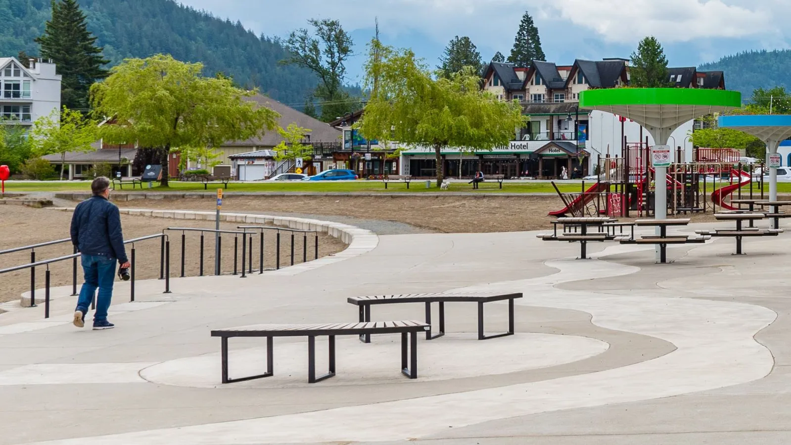 Skyline benches provide seating along a concrete walkway near a playground and beach. The benches feature recycled plastic lumber and a black powder-coated frame, complementing the recreational area.