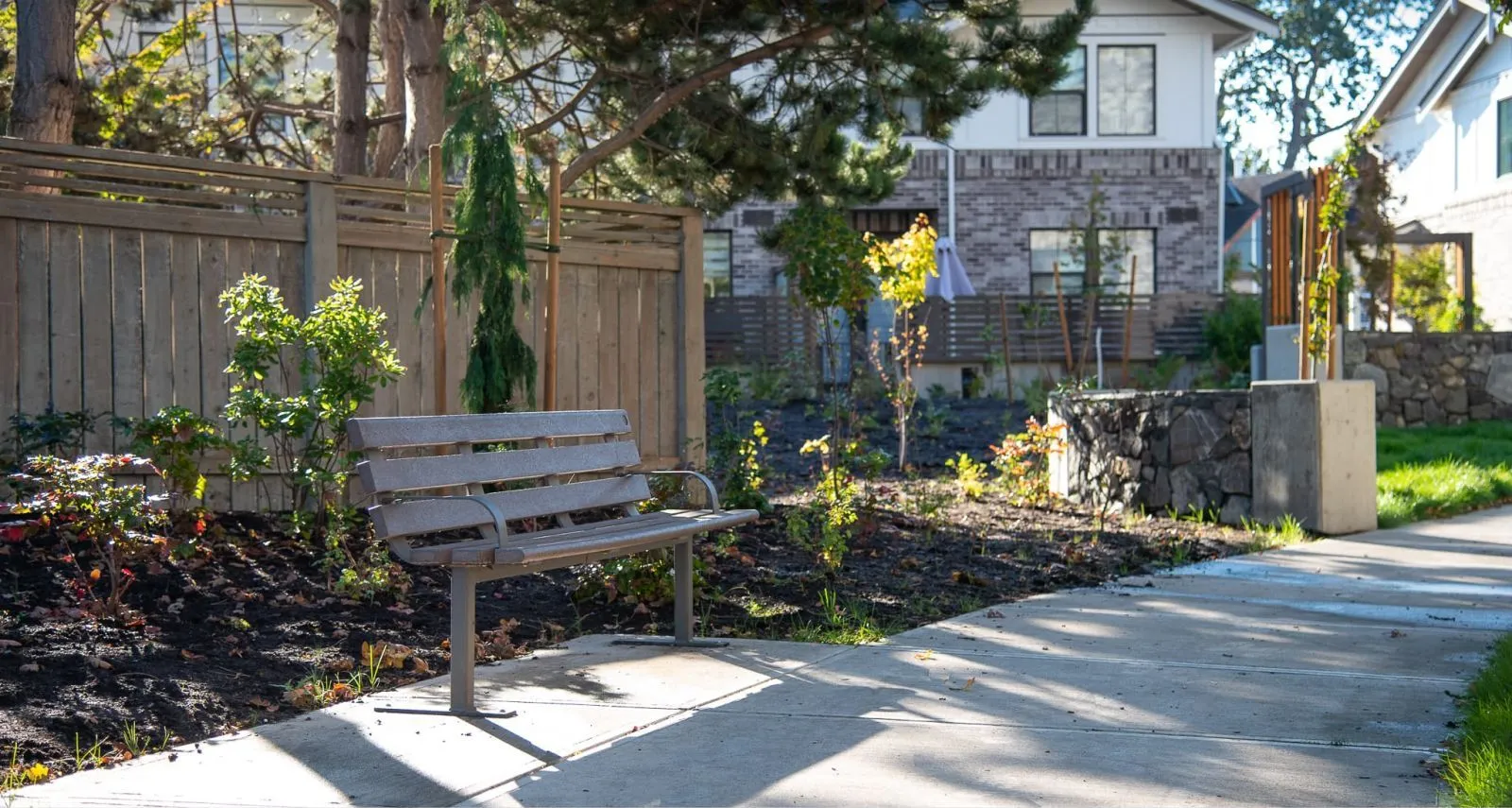 A Standard Bench with Armrests provides seating along a sidewalk in a residential neighborhood. The bench features recycled plastic lumber slats and a powder-coated metal frame.