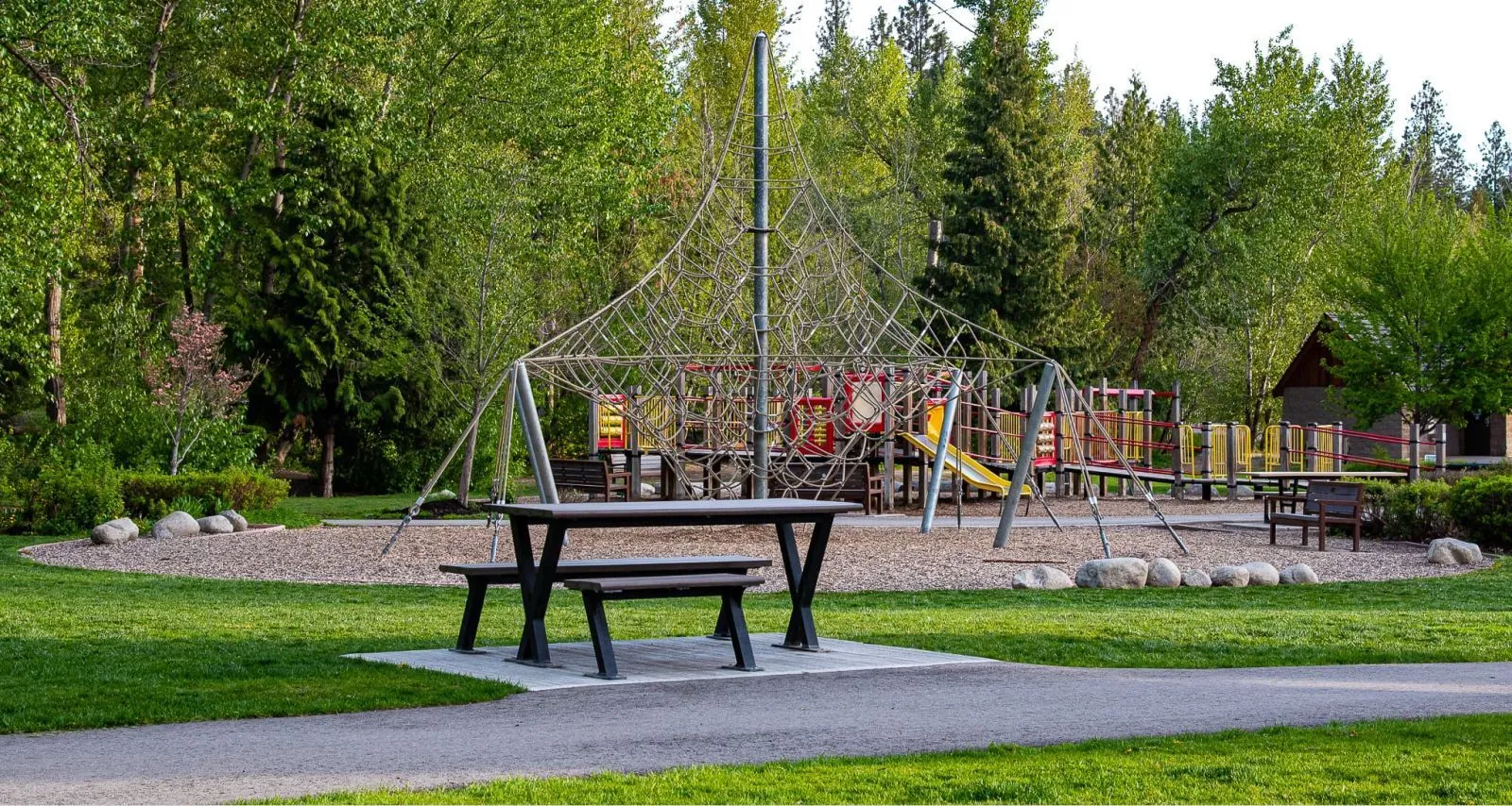 Wheelchair accessible picnic table in a park setting. The table features a black frame and is surrounded by lush greenery and a playground.