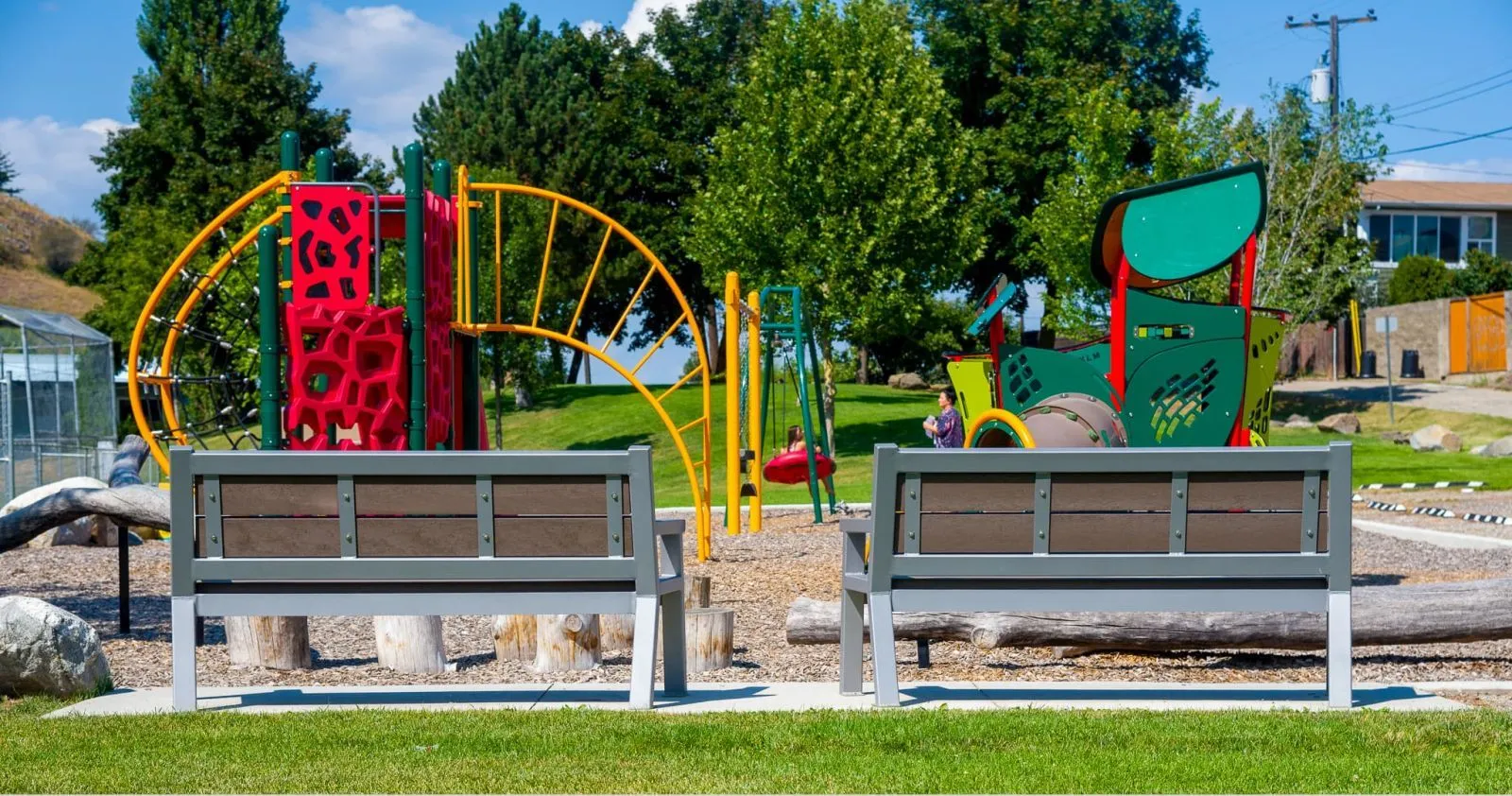 Two Rutherford Wide Body benches face the playground at Alexis Park in Vernon, British Columbia. The benches feature recycled plastic lumber and powder-coated metal frames.