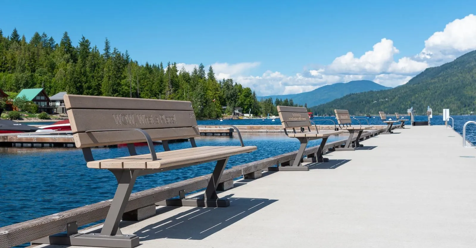 Row of Bayview benches line a pier overlooking a lake surrounded by forested mountains. These durable benches offer comfortable seating and scenic views.