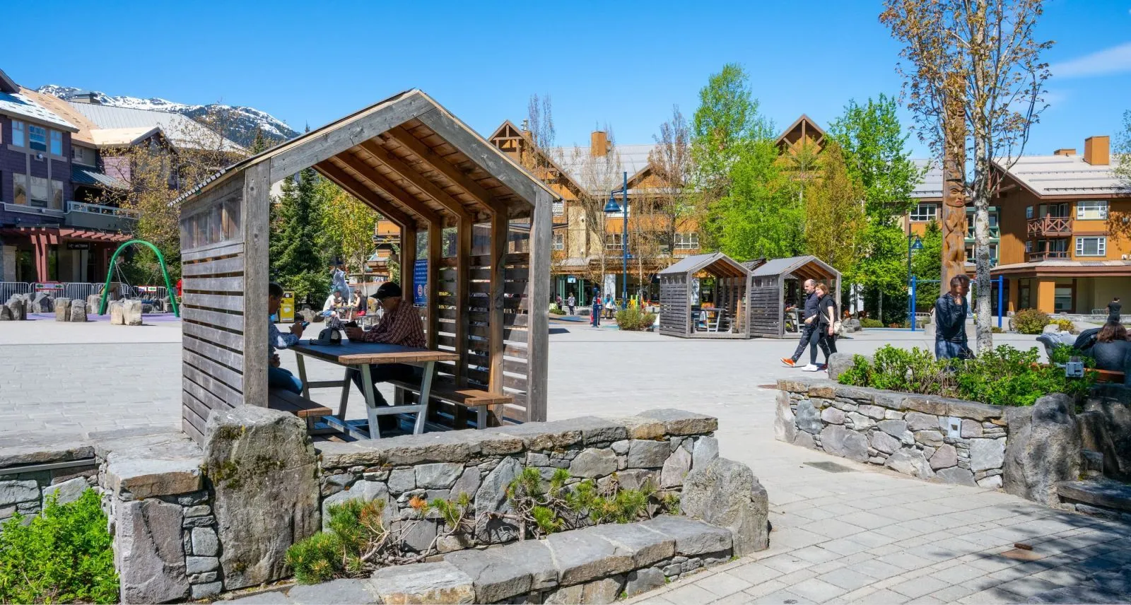 Heavy-duty picnic tables provide a gathering space in a public square. The tables are sheltered by wooden structures, offering a comfortable spot for people to relax and enjoy the surroundings.
