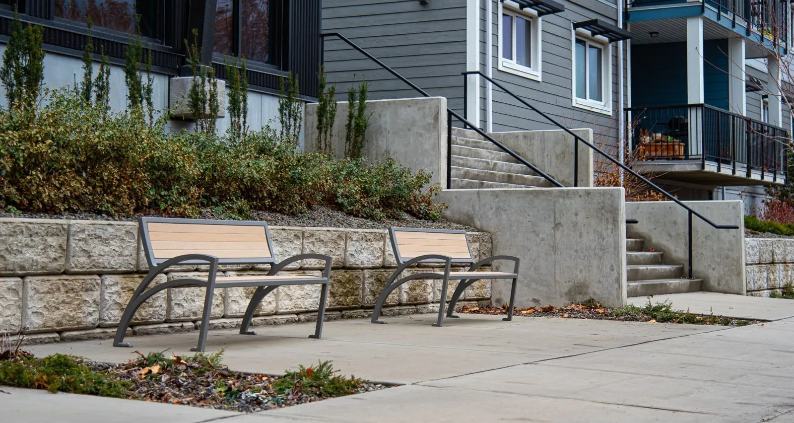Two Modena benches provide seating outside a modern apartment building. The benches feature recycled plastic lumber seats and powder-coated metal frames.