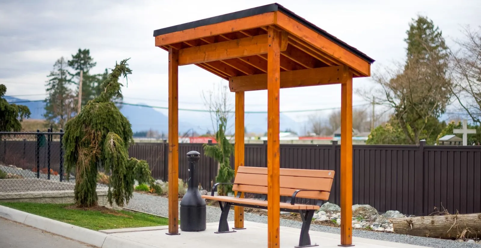 Sheltered bench seating area with a Parker Bench and black litter bin. This outdoor furniture provides a comfortable and functional space in a landscaped area.