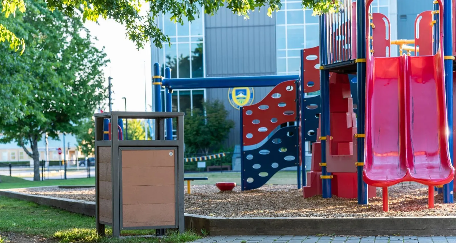 Urban Form Easy Access Waste Receptacle at a playground. The bin features a brown recycled plastic lumber body and a gun metal powder-coated frame.
