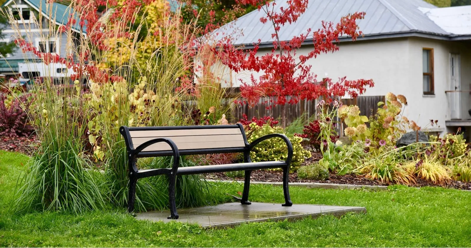 A Mountain Classic bench sits in a garden setting with colorful foliage. The bench features recycled plastic lumber slats and a powder-coated metal frame.