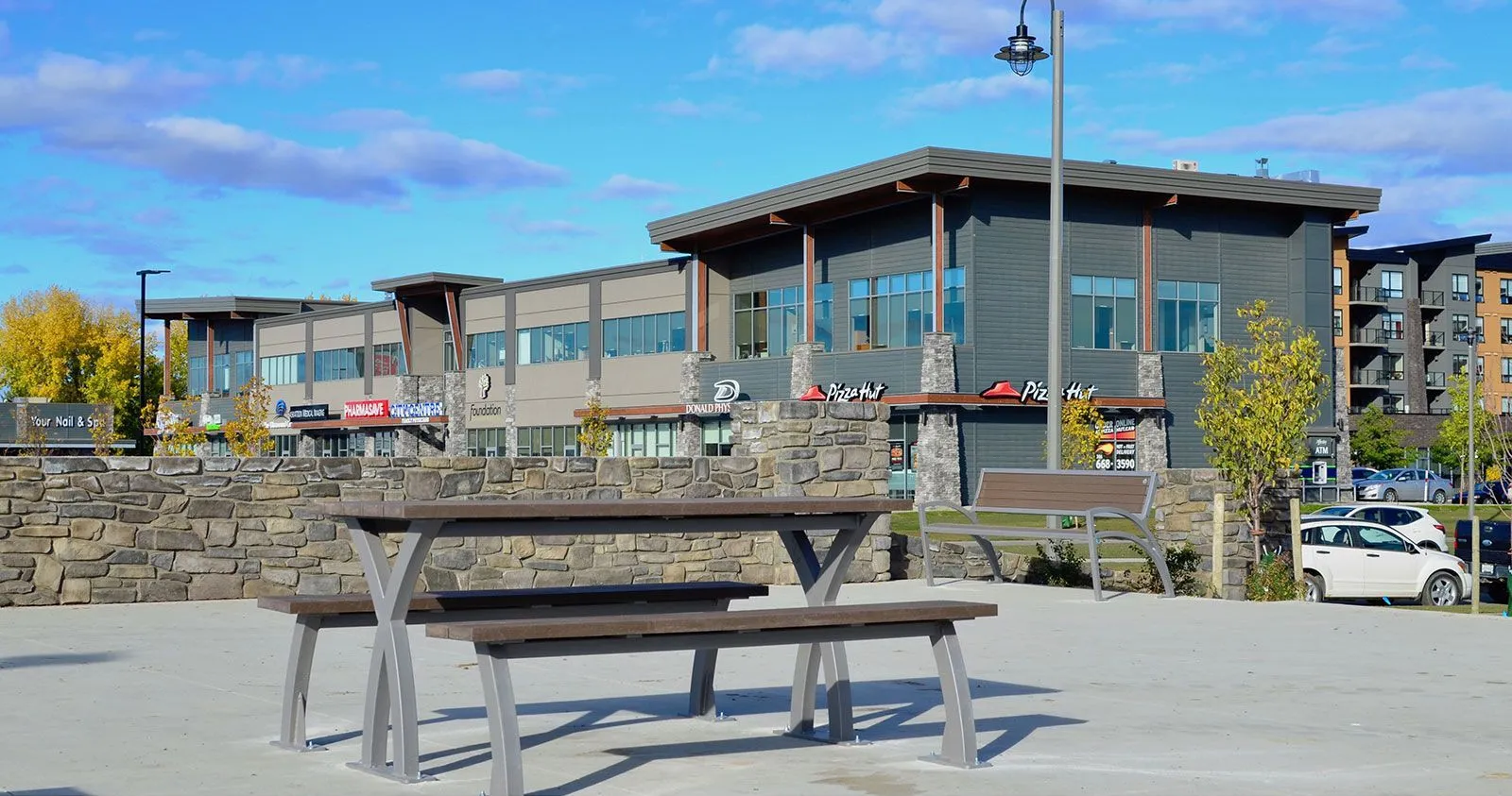 This outdoor space features a Parker picnic table and bench seating, providing a comfortable area for gatherings. The setting includes commercial buildings and stone retaining walls.