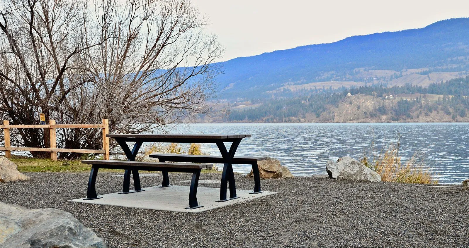 A picnic table with benches sits on a gravel shore next to a lake, with mountains in the background. The table features recycled plastic lumber and a black powder-coated frame.