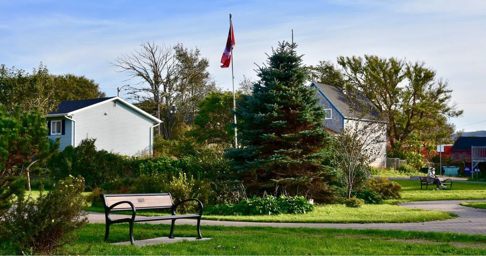 A Mountain Classic Wide Body Bench sits in a park-like setting with a Canadian flag in the background. This bench provides comfortable seating in outdoor spaces.