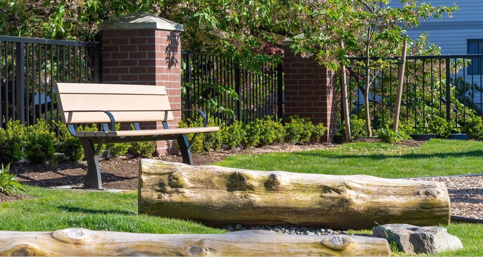 A Parker bench provides seating in a landscaped area with a brick and iron fence. The bench features recycled plastic lumber and a powder-coated metal frame.