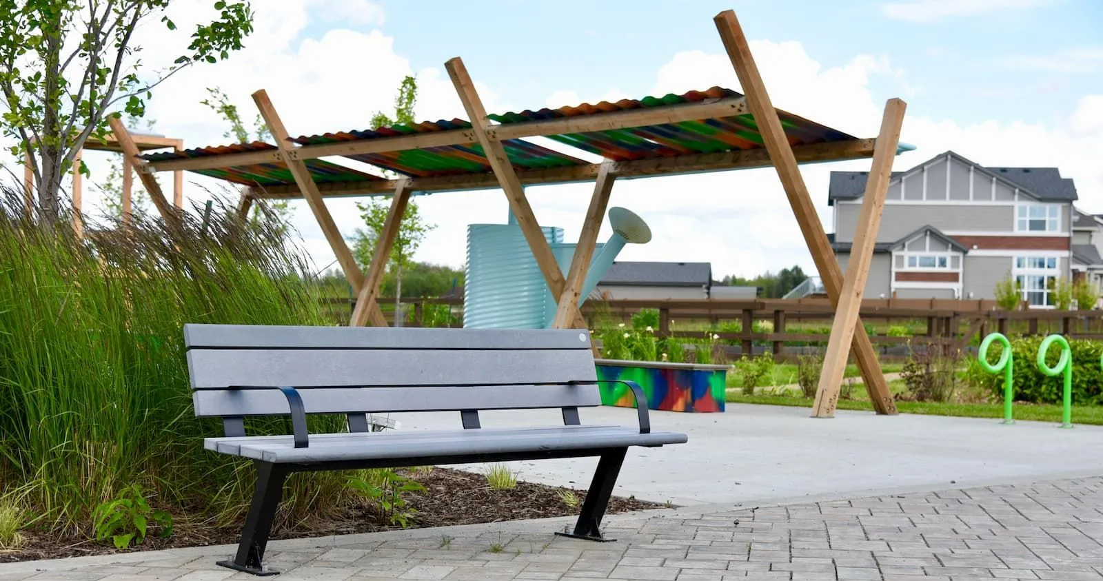 A Bayview bench with black powder-coated metal frame and grey recycled plastic lumber seat is installed in a park setting. The bench sits on a paved path near a wooden pergola and landscaping.