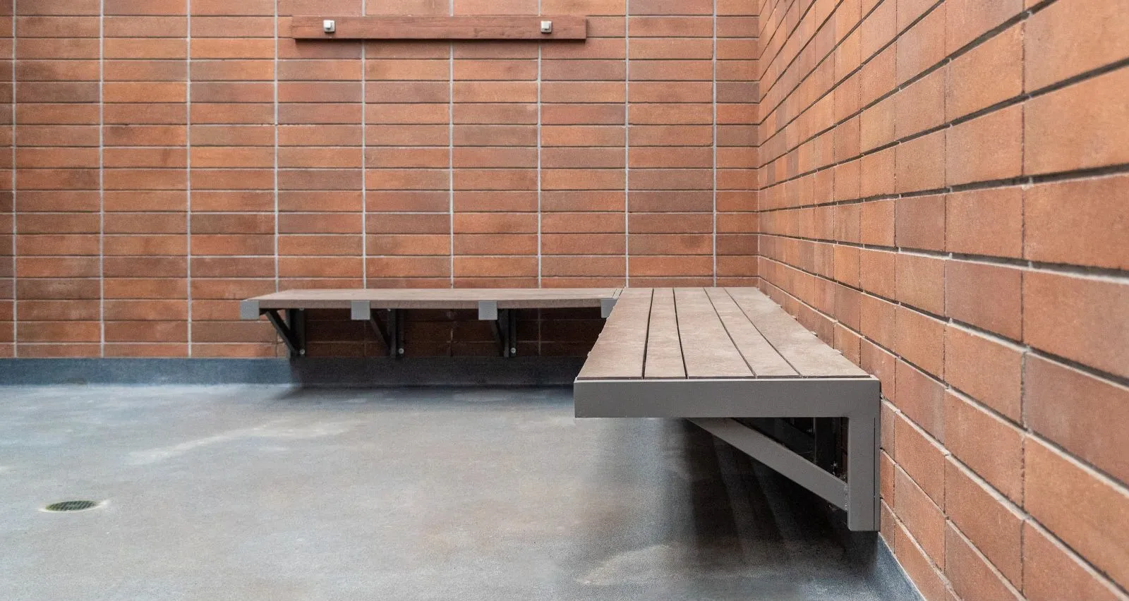 Wall-mounted benches offer seating in a brick-walled alcove, providing a functional and stylish solution for public spaces. The benches feature recycled plastic lumber and powder-coated metal frames.