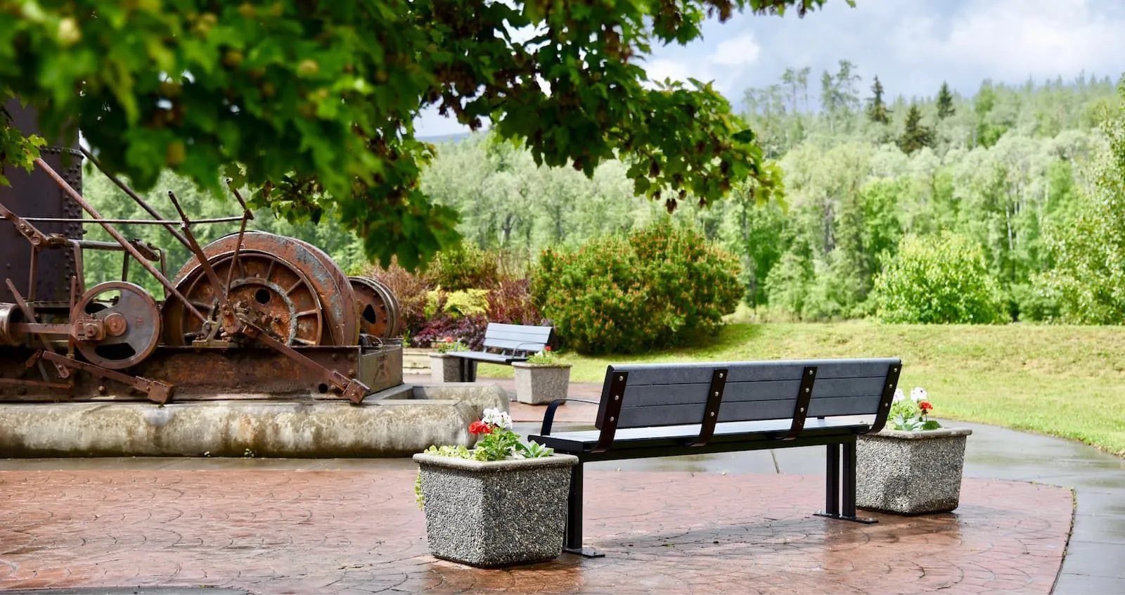 A Larson bench sits on a brick-paved area next to an old mining artifact. The bench features recycled plastic lumber and a powder-coated metal frame, providing a comfortable resting spot in the park.