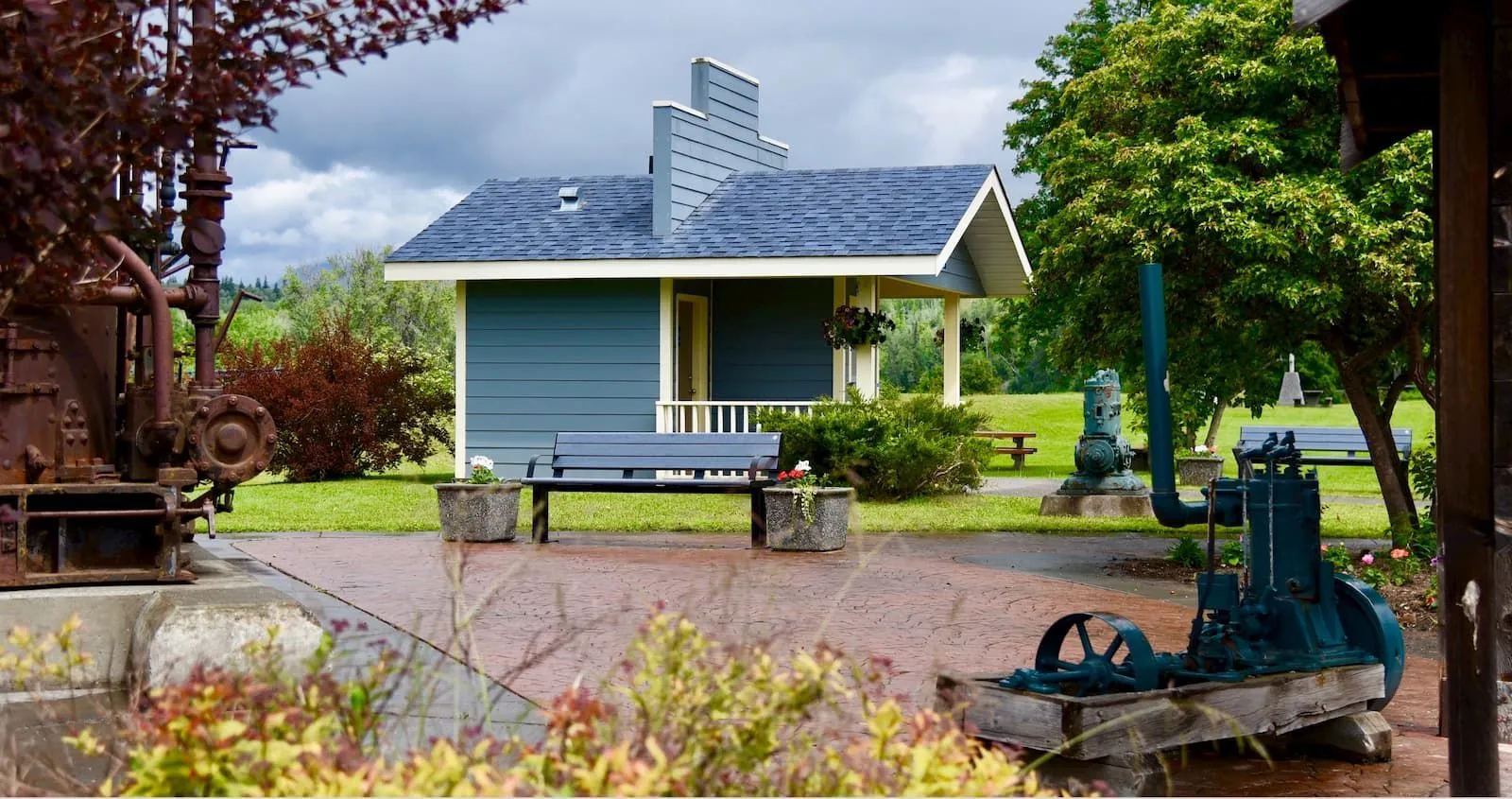 A Larson Bench provides seating in a park-like setting with vintage machinery and a small building. The bench offers a place to rest and enjoy the surrounding scenery.