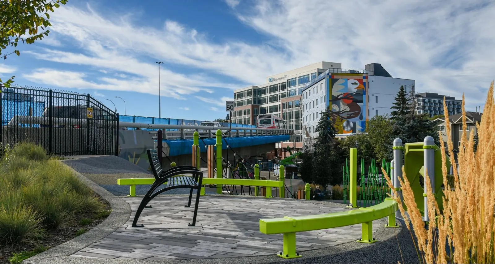 A black Modena Aluminum Bench sits in Flyover Park, Calgary, Alberta. The park features a playground and public art, with a bridge in the background.