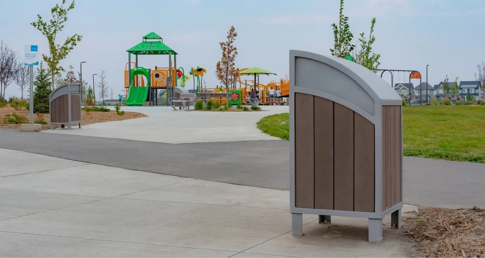Curved top waste receptacles are placed in a park setting near a playground. The bins feature recycled plastic lumber and powder-coated metal frames.