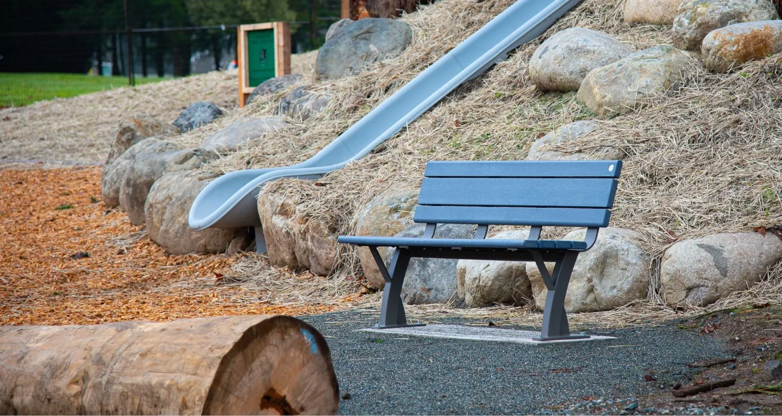 A Parker bench sits near a slide in a park setting. The bench features recycled plastic lumber and a powder-coated metal frame.