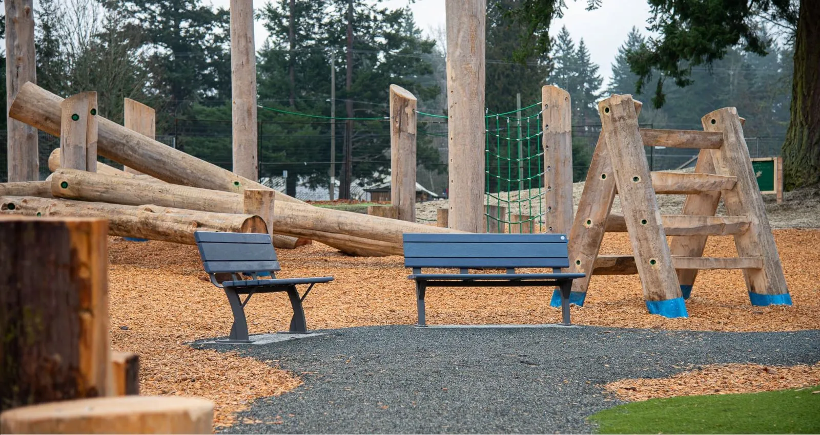 Two Parker benches provide seating at Elk/Beaver Lake Regional Park in Saanich, British Columbia. The benches are situated near a wooden playground structure.