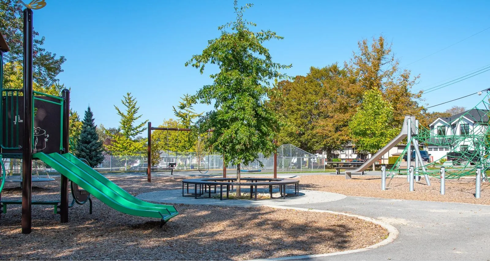 A circular bench surrounds a tree in a playground setting. The park also features slides, swings, and picnic tables.