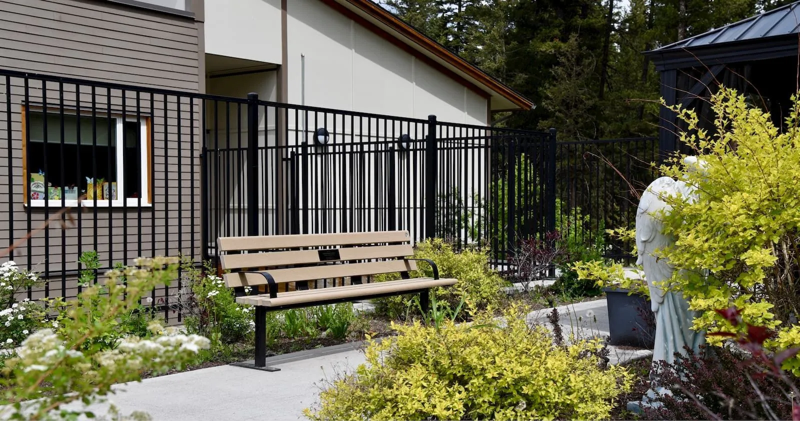 A standard memorial bench with armrests provides seating in a landscaped garden setting. The bench features recycled plastic lumber and a black powder-coated frame.