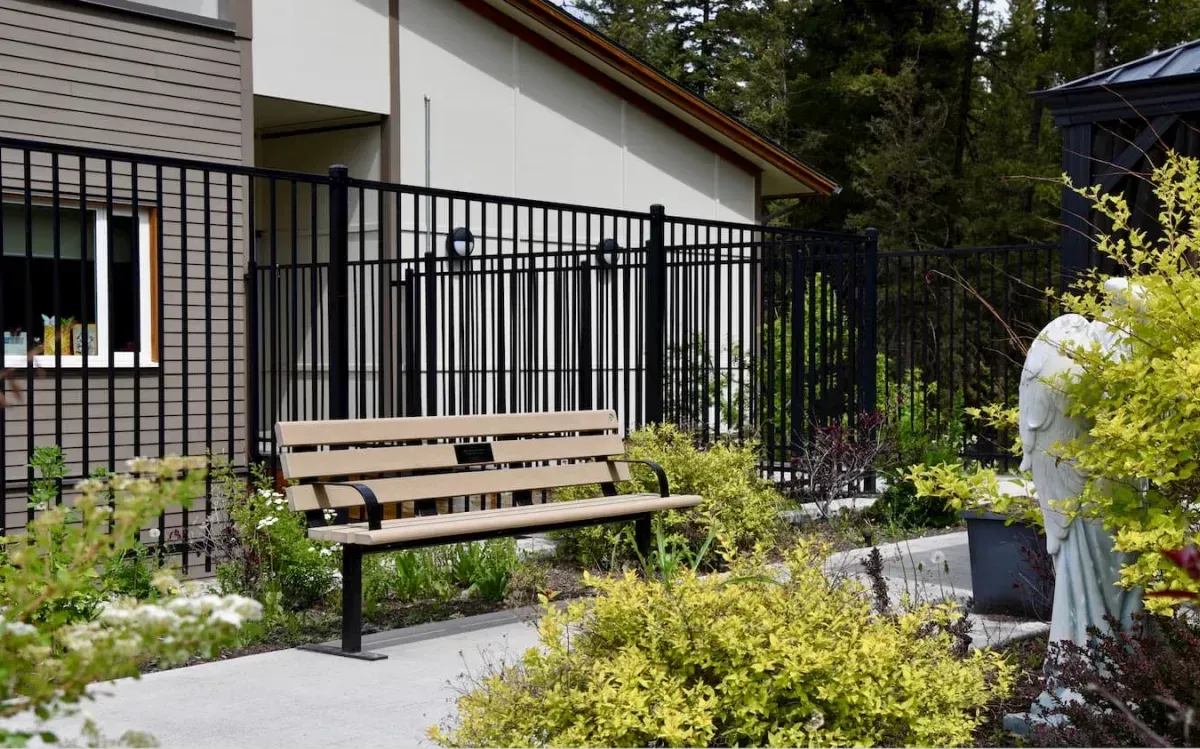 A standard memorial bench with armrests provides seating in a landscaped garden area. The bench features recycled plastic lumber slats and a black powder-coated frame.
