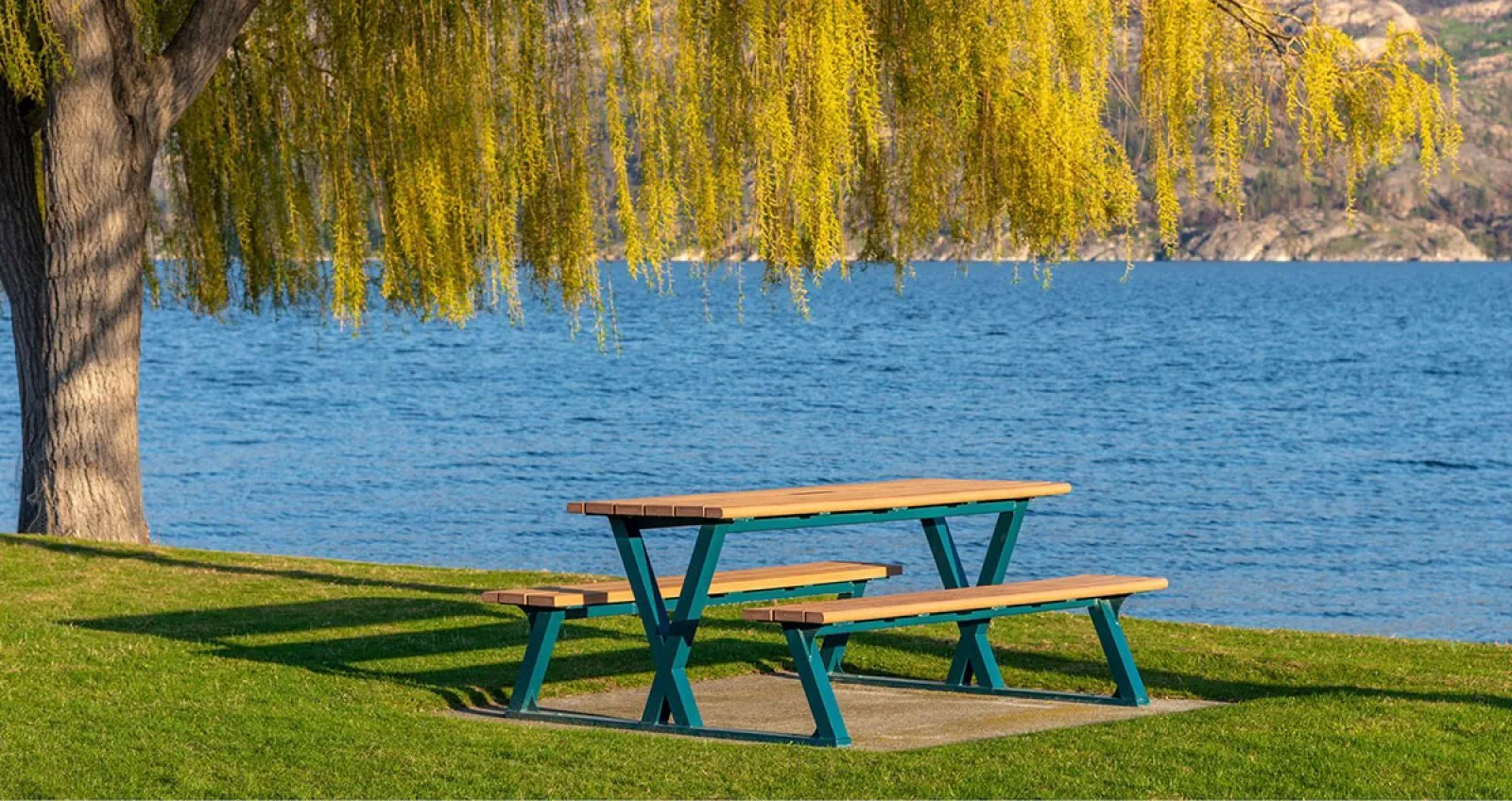 A Bayview picnic table sits beside a lake under a weeping willow tree. The table features recycled plastic lumber and a powder-coated metal frame.