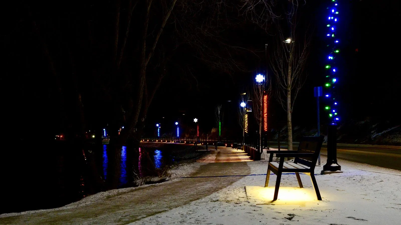 A Rutherford bench sits along a snow-covered path illuminated by festive lights at night. The bench provides a place to rest and enjoy the winter scenery.