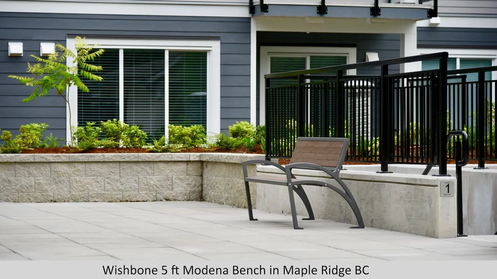 A Modena bench provides seating in an outdoor space. The bench features recycled plastic lumber and a powder-coated metal frame, complementing the modern building and landscaping.