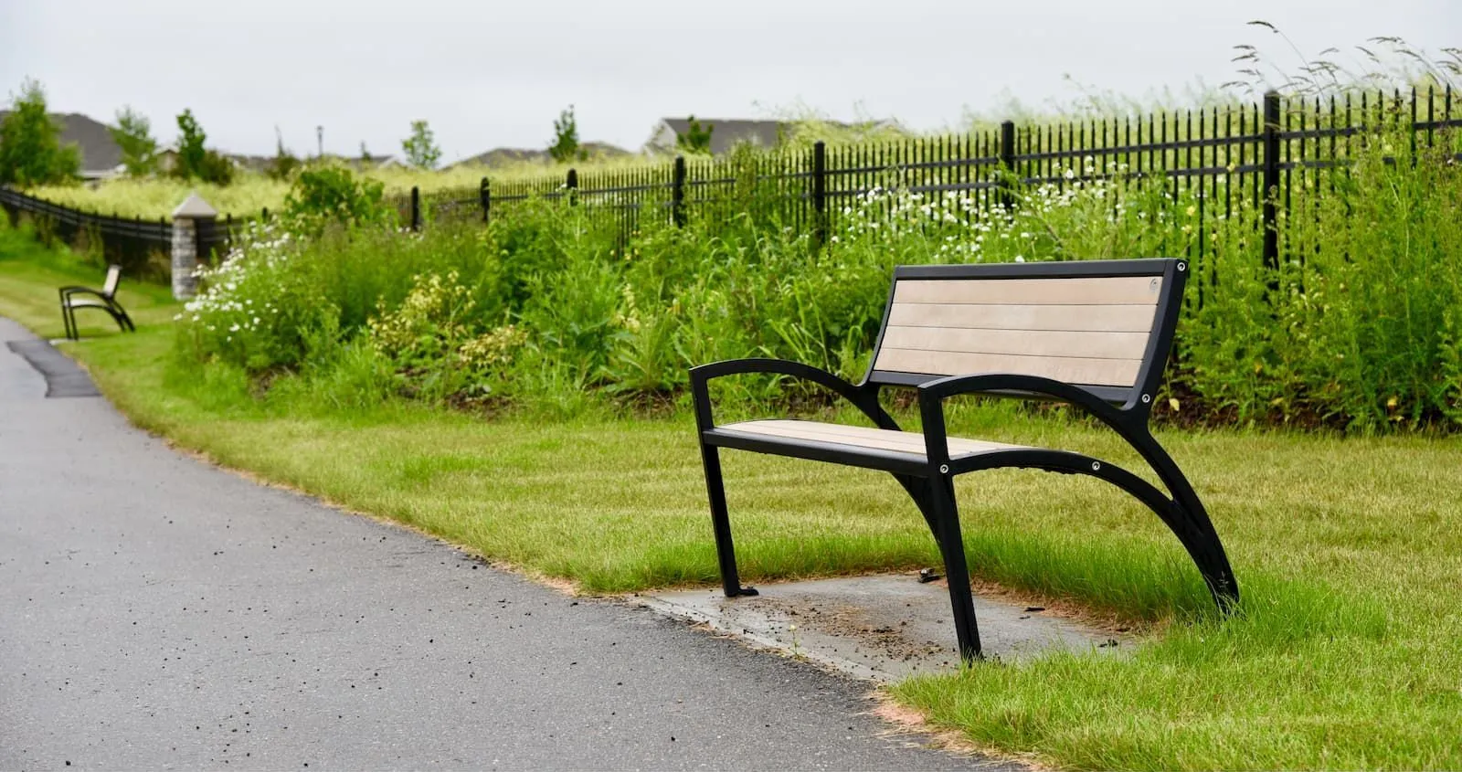 A Modena bench sits along a paved path in a grassy area. The bench features recycled plastic lumber slats and a black powder-coated frame.