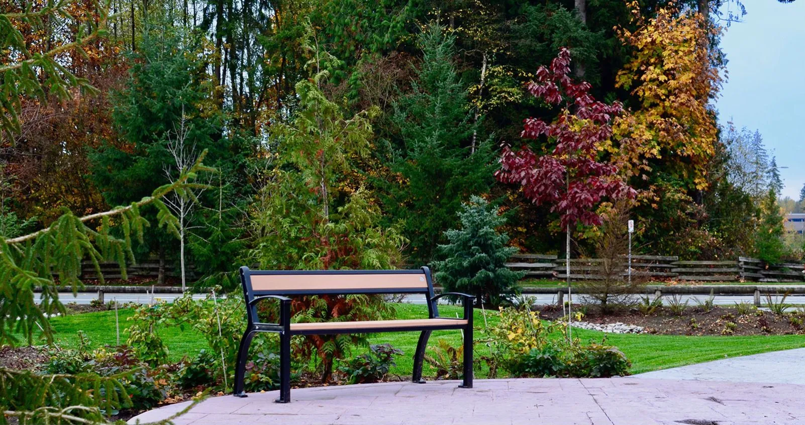 A Beselt bench provides seating in a landscaped park setting. The bench features recycled plastic lumber slats and a powder-coated metal frame.