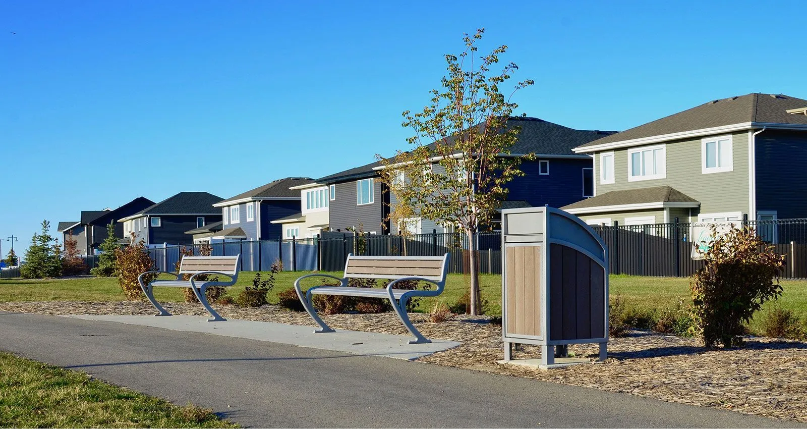 Two Modena benches and a Modena waste receptacle sit along a paved path in a residential area. The site furniture provides a place to rest and dispose of waste along the walking trail.