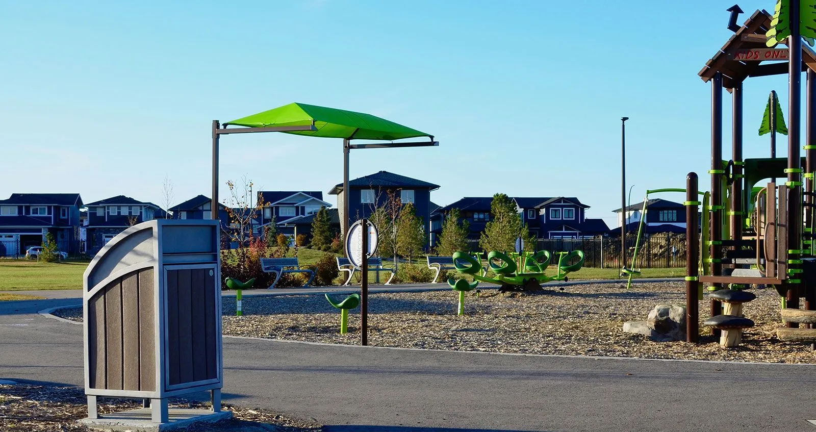 Modena curved top waste receptacle stands near a playground in a residential area. This litter bin provides a convenient disposal solution for public spaces.