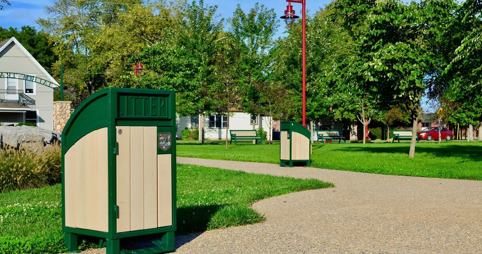 Curved top waste receptacles are placed along a paved path in a park setting. The bins feature a green frame and light-colored recycled plastic lumber.