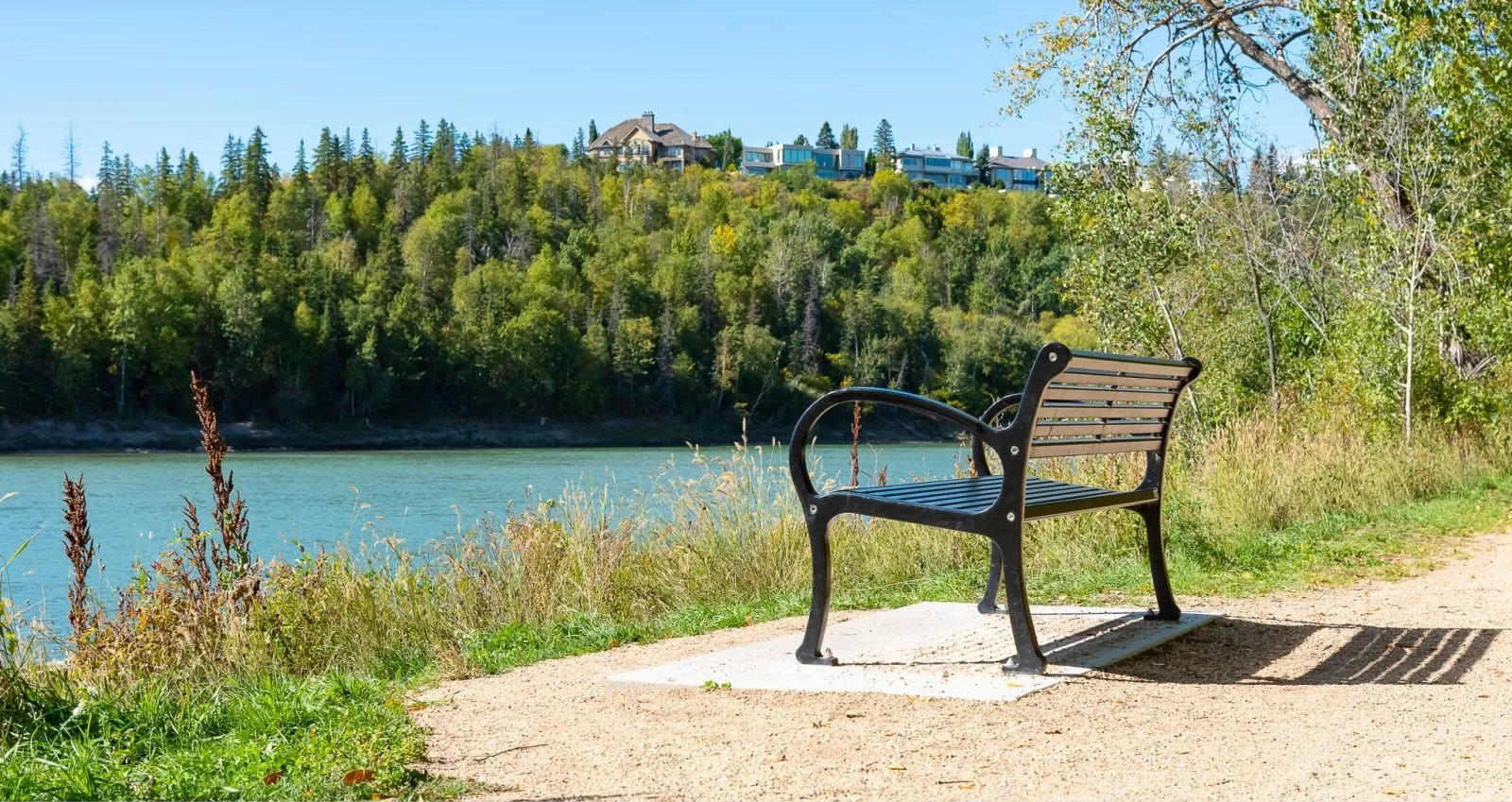 A Mountain Classic bench sits along a riverside path in a park setting. The bench features a black frame and light-colored recycled plastic lumber.