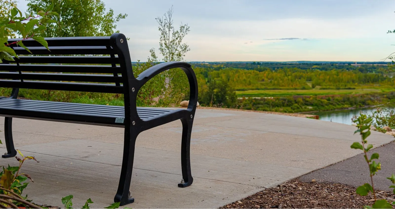 A black Mountain Classic bench offers a place to rest and enjoy the view of the River Valley Initiative East Trail in Edmonton, Alberta. The bench sits on a concrete pad overlooking a lush green valley and river.