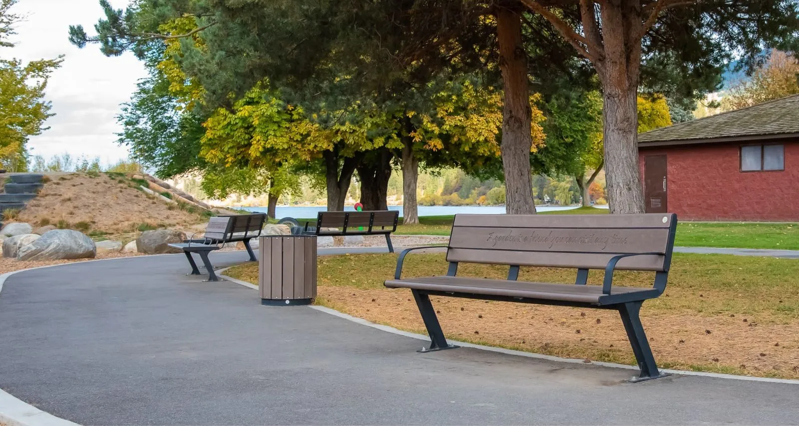Bayview Plus benches line a paved path in Beasley Park, offering a place to rest and enjoy the lakeside views. A matching waste receptacle complements the site furniture.