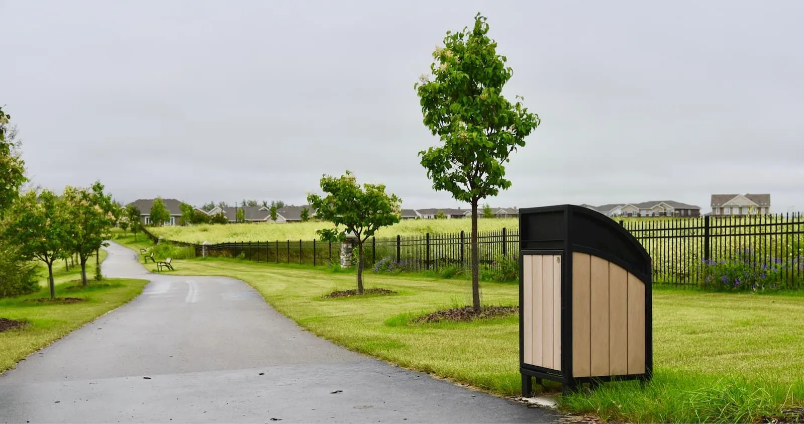 Modena Curved Top Waste Receptacle in a park setting. The bin features recycled plastic lumber and a black powder-coated frame.