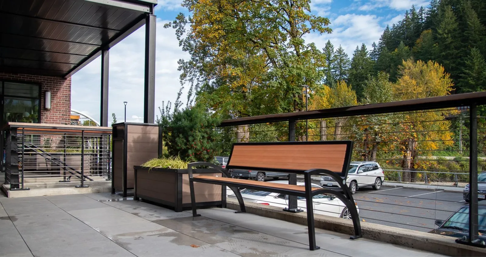 This outdoor space features a Modena Wide Body Bench with recycled plastic lumber and a black powder-coated frame, accompanied by a planter box. The bench offers seating with a view of the parking area and surrounding trees in Chilliwack, BC.