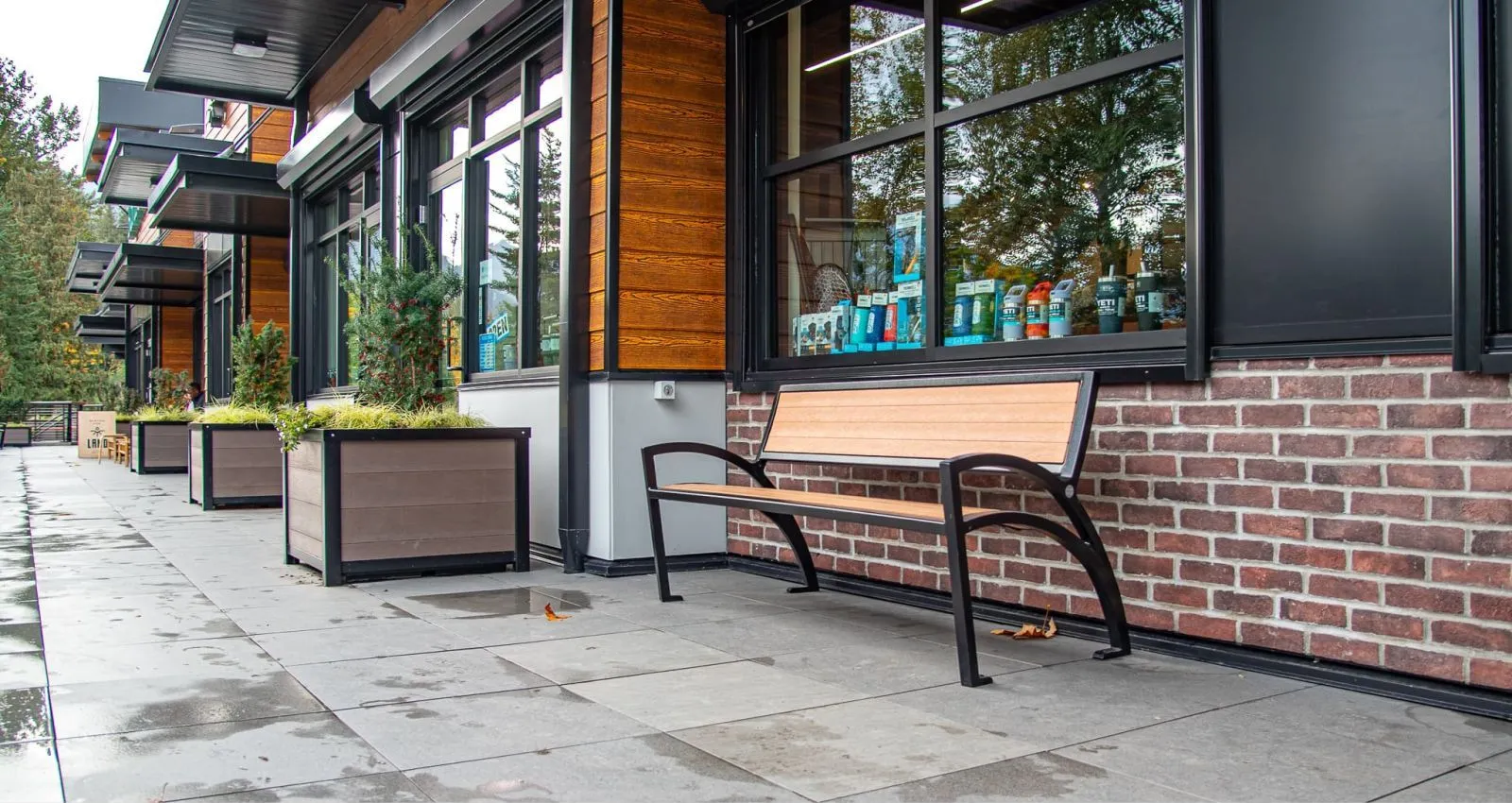 A Modena Wide Body Bench provides seating outside a commercial building. The bench features recycled plastic lumber slats and a powder-coated metal frame.