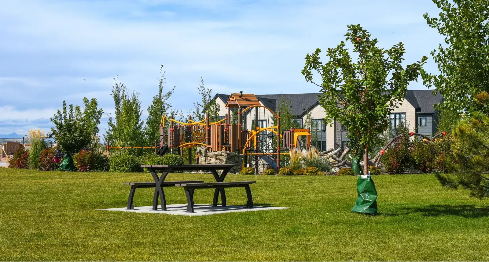 A Parker Picnic Table sits on a grassy lawn in a community park. The table features a black frame and a natural-looking top, providing a comfortable space for outdoor dining and relaxation.