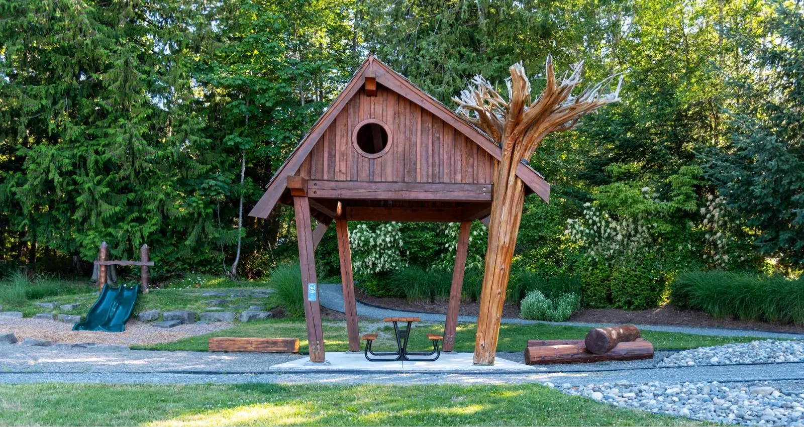 A black Pipeline Picnic Table sits under a wooden shelter in a park-like setting. The table provides a place for visitors to rest and enjoy the surrounding nature.