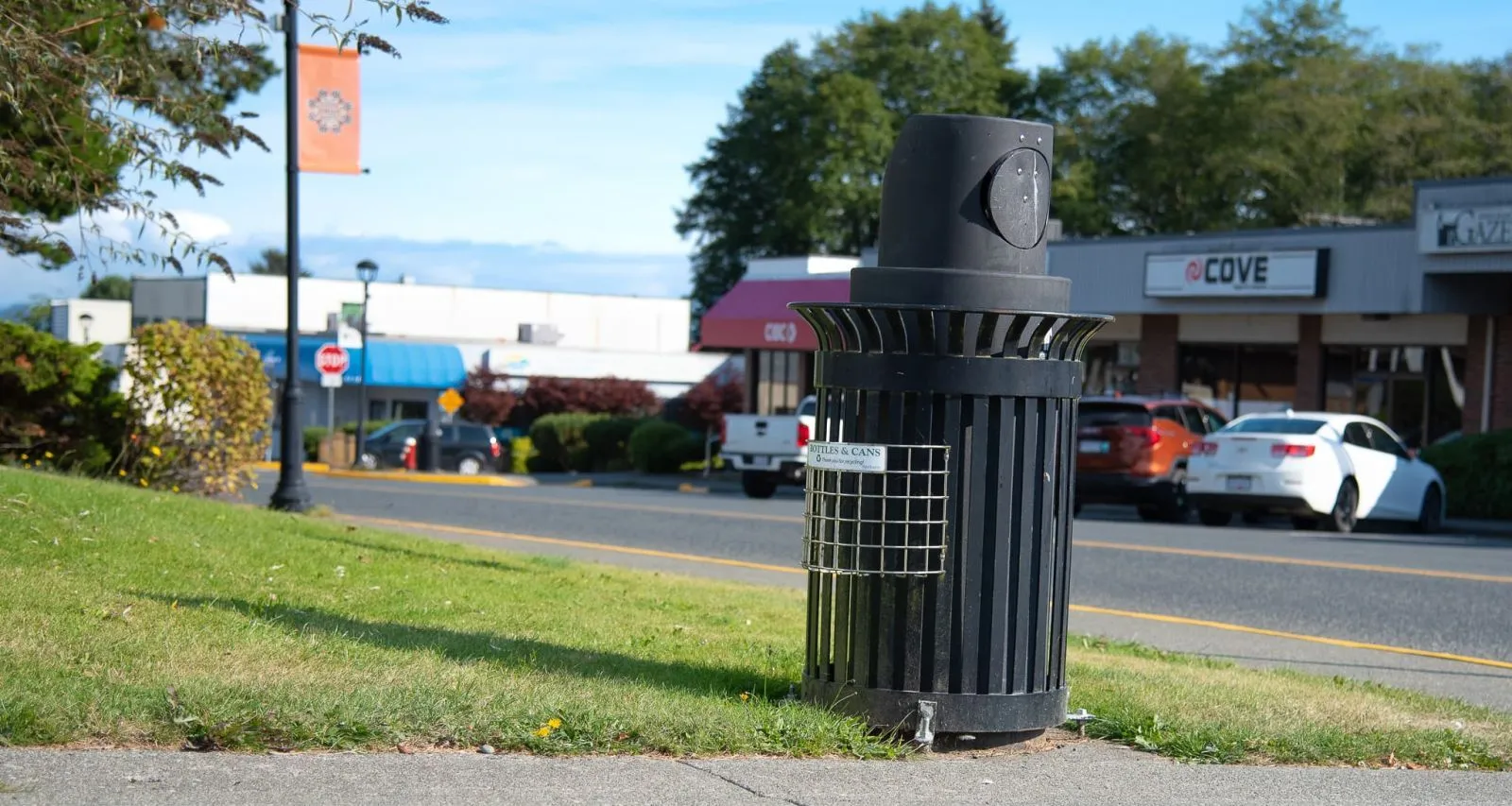 A black round bottle and can recycling basket is located in Port Hardy, British Columbia. The bin is situated on a sidewalk next to a grassy area, with commercial buildings visible in the background.