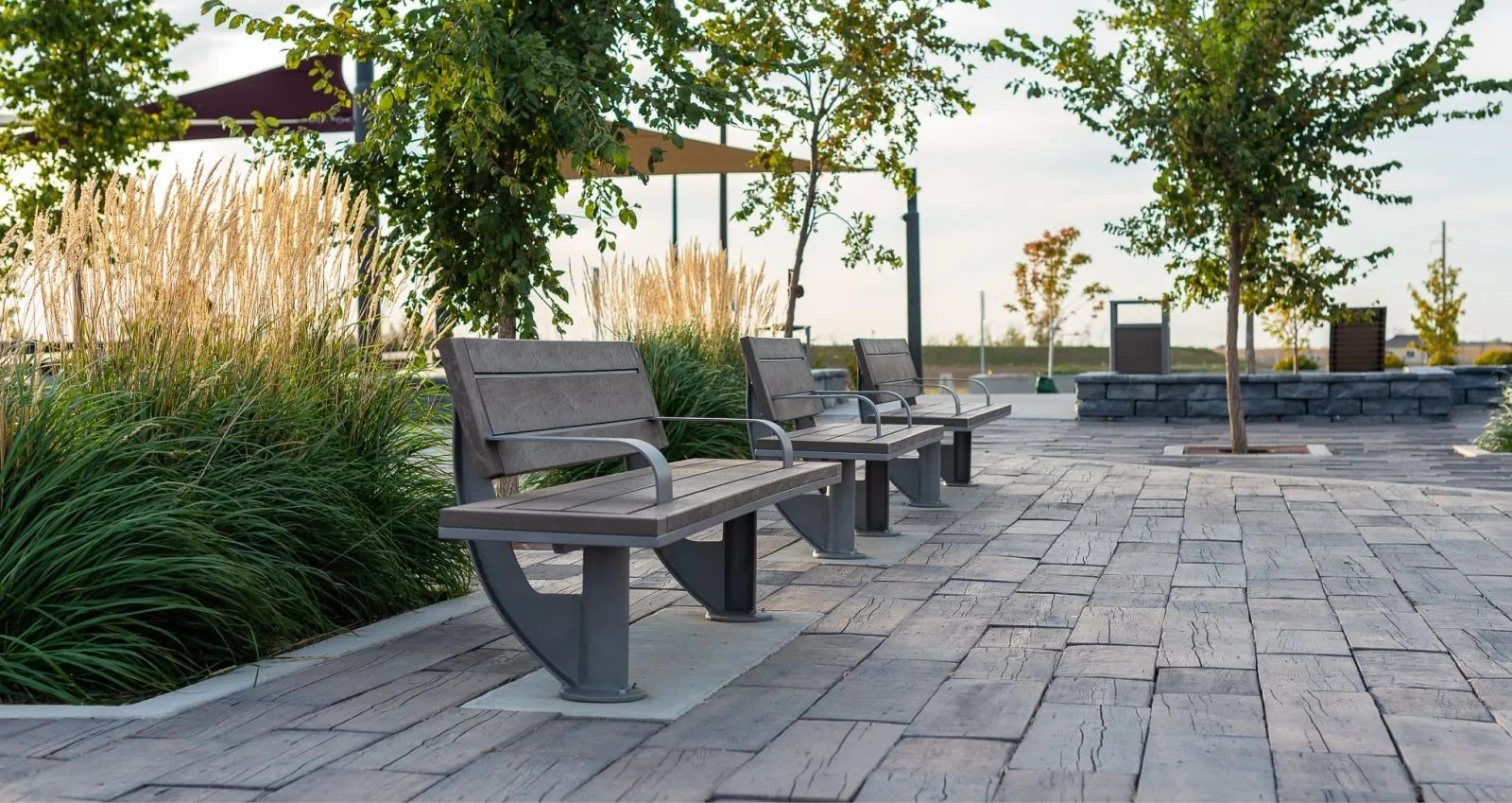 Three SurRe benches line a paved walkway in a landscaped park. The benches feature recycled plastic lumber seating and powder-coated metal frames.