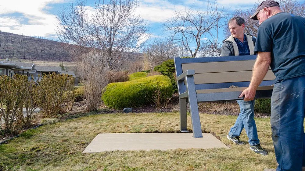 A bench is being installed on a concrete pad in a grassy area with trees and buildings in the background. The bench features recycled plastic lumber and a powder-coated metal frame.