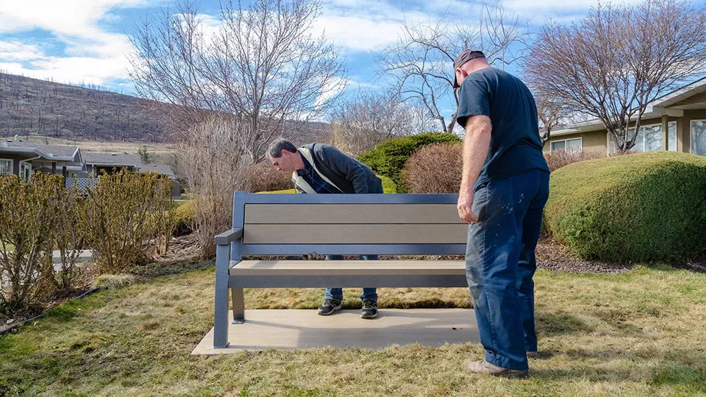A durable bench with recycled plastic lumber seating and a powder-coated metal frame provides comfortable outdoor seating. This bench is installed on a concrete pad in a landscaped area.