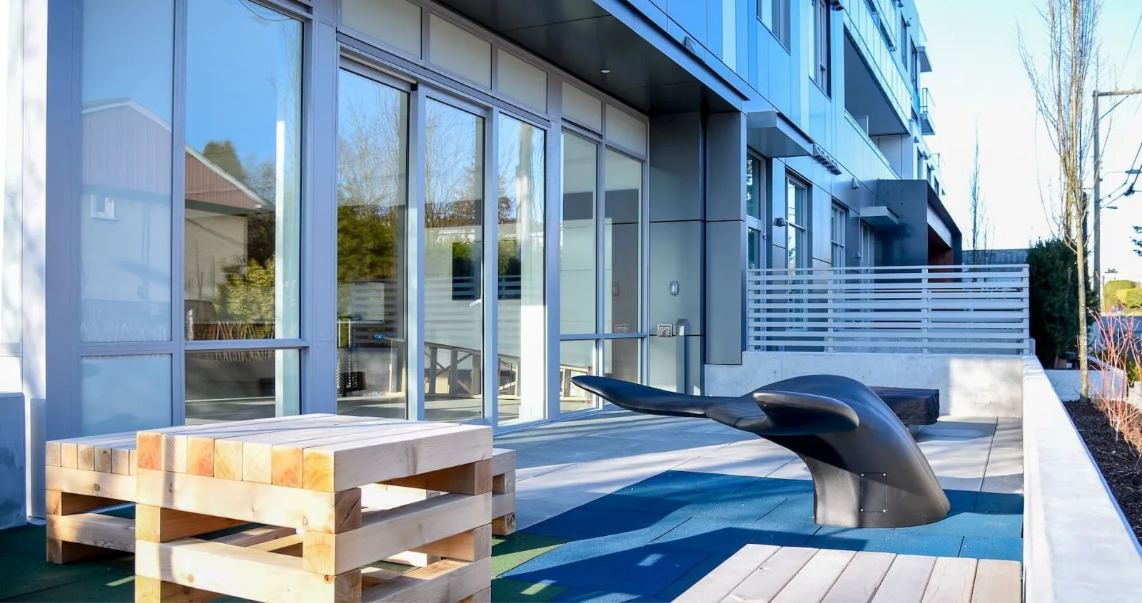 Aluminum Whale Tail bench provides a unique seating option in an outdoor space. The bench is paired with wooden pallet-style benches in front of a modern building.